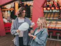 Ein Paar teilt sich Zuckerwatte vor einem "Roll a Ball"-Spiel im Freizeitpark. Ihr lässiger Freizeitpark-Outfit passt perfekt zum Tag.