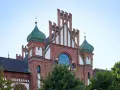 Das imposante Gebäude im Stil des Zollern-Schlosses mit seinen grünen Kuppeln und roten Ziegeln ragt vor blauem Himmel auf.