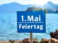Schild am Strand: "1. Mai Feiertag". Die Sonne scheint über dem Meer, ideal für Veranstaltungen am 1. Mai.