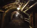 Dunkler Stollen unter Tage mit Gleisen und Rohren, beleuchtet von Lampen.