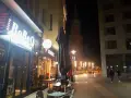 Ein beleuchtetes Restaurant "BoBoQ" und eine Kirche prägen das Bild von Dortmund bei Nacht. Menschen spazieren auf dem Gehweg, während Bauarbeiten im Hintergrund stattfinden.
