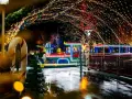 Ein bunter Zug fährt durch eine festlich beleuchtete Gasse im winterwonder legoland. Lichterketten und Kugeln schmücken den Weg.