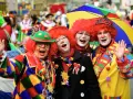 Vier Clowns mit roten Perücken und bunter Kleidung lachen und winken. Ein bunter Regenschirm ist über ihnen. Hier wird kein Karneval gefeiert, sondern ausgelassen gefeiert!