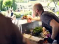 Mann reicht ein Gericht auf einem Holzbrett bei einem Street Food Festival. Die Preise sind hier fair für die leckere Auswahl.