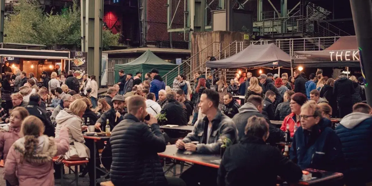 Menschen genießen Speisen und Getränke auf dem Street Food Festival Duisburg. Viele Besucher sitzen an langen Tischen und unter Zelten.