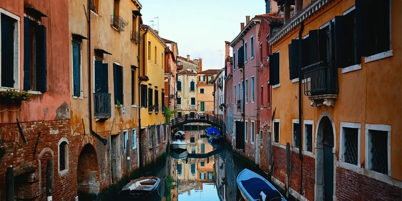 Bunte Häuser säumen einen Kanal in Venedig. Kleine Boote treiben auf dem Wasser. Ein kleiner Bogenbrücke überspannt den Kanal. Das ist, was man in Venedig gesehen haben muss.