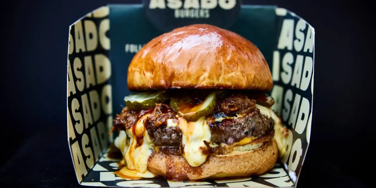 Saftiger Burger mit geschmolzenem Käse und Gurken, präsentiert in einer "ASADO BURGERS" Box. Der beste Burger Kölns wartet!
