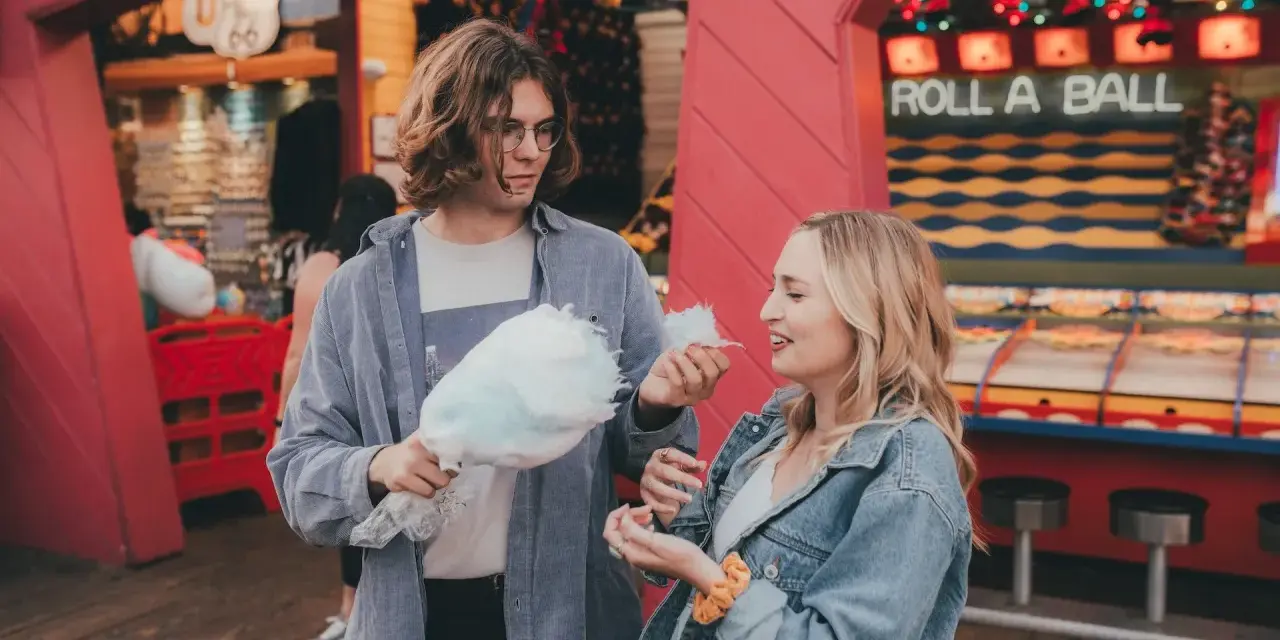Ein Paar teilt sich Zuckerwatte vor einem "Roll a Ball"-Spiel im Freizeitpark. Ihr lässiger Freizeitpark-Outfit passt perfekt zum Tag.