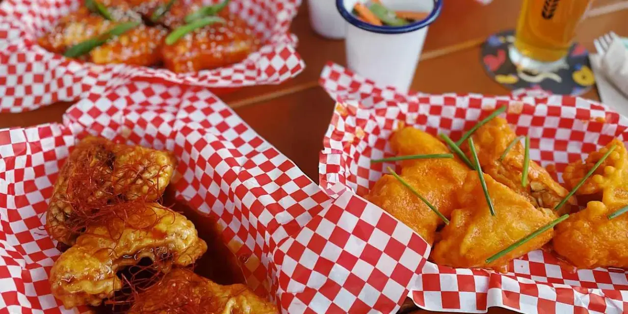 Leckere Cosmo Wings in roten Körbchen, garniert mit Chilis und Schnittlauch. Dazu ein Bier und kleine Beilagen.