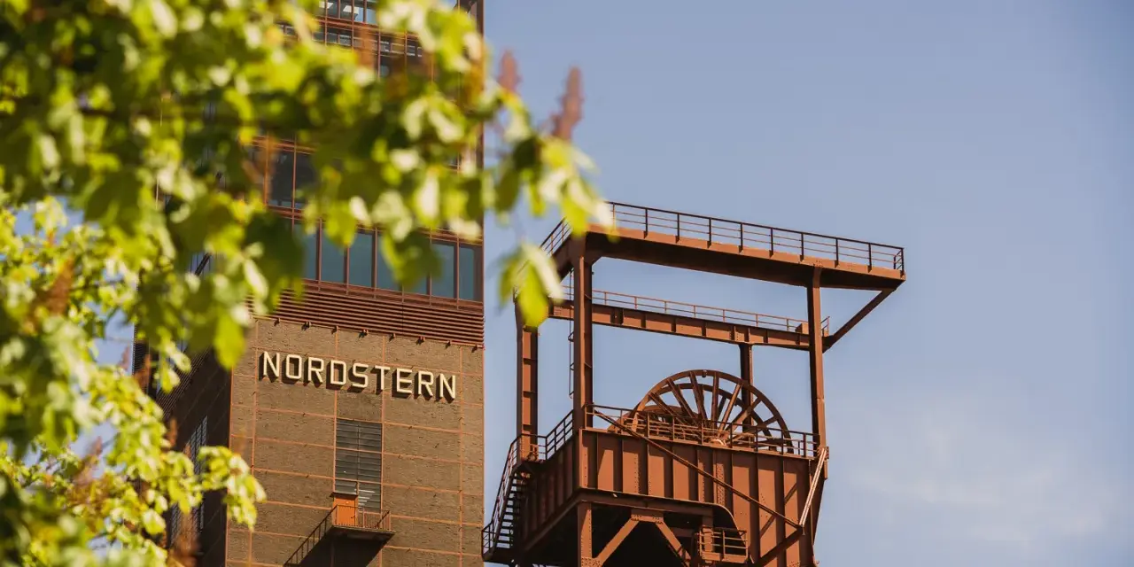 Industriearchitektur im Nordsternpark Gelsenkirchen: Ein markanter Förderturm und das Gebäude mit der Aufschrift "NORDSTERN" vor blauem Himmel.