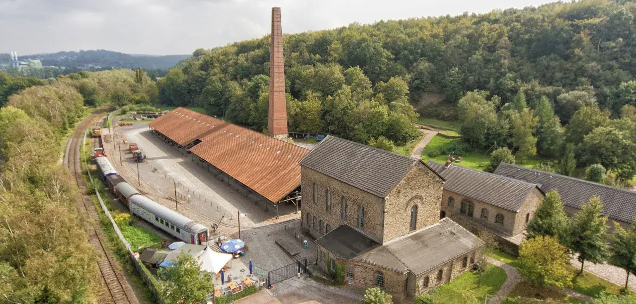 Alte Zechengebäude der Zeche Nachtigall mit einem langen roten Dach, einem hohen Backsteinschornstein und einem historischen Zug.
