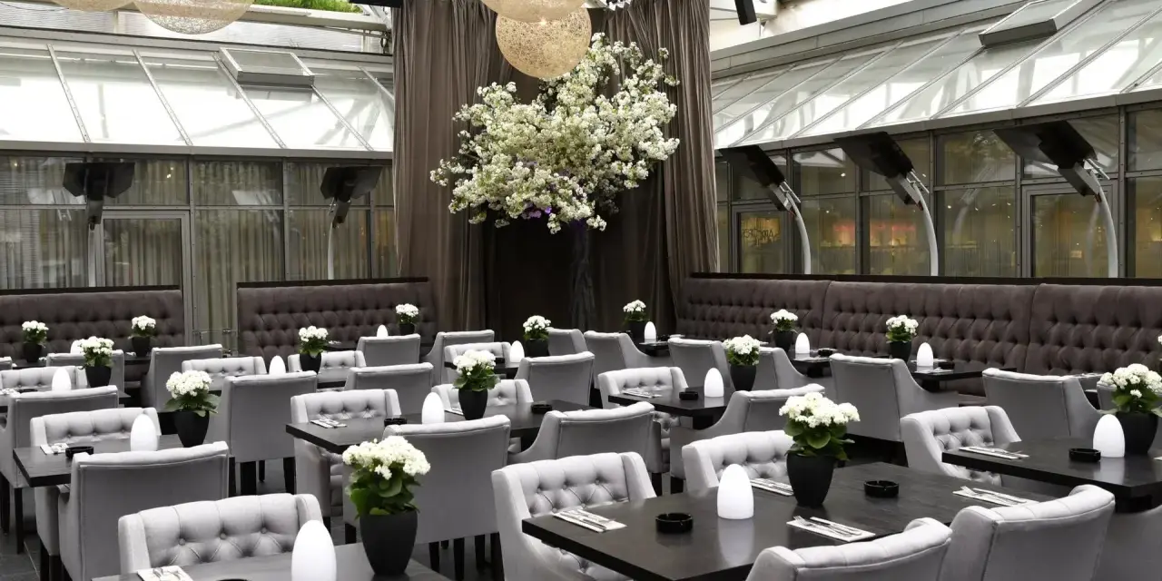 Elegantes Restaurant mit grauen Sitzmöbeln und dunklen Tischen, dekoriert mit weißen Blumen. Ein großer Kirschblütenbaum im Zentrum, beleuchtet von kugelförmigen Lampen. Perfekt für ein Rendezvous, wie ein Oscar-Moment im Apropos.