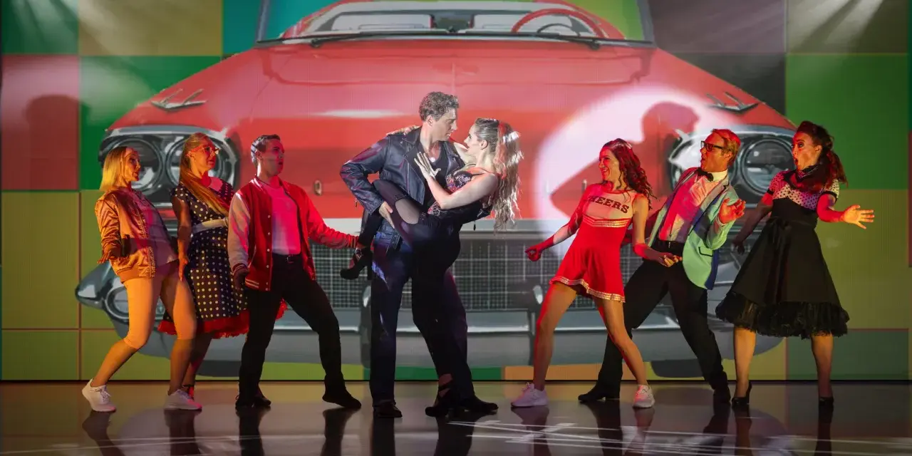 Tänzer in 50er-Jahre-Outfits vor einem roten Oldtimer-Cabriolet. Ein Paar tanzt im Zentrum, umgeben von anderen Darstellern. Ein lebendiger Moment aus Musicals Deutschland.