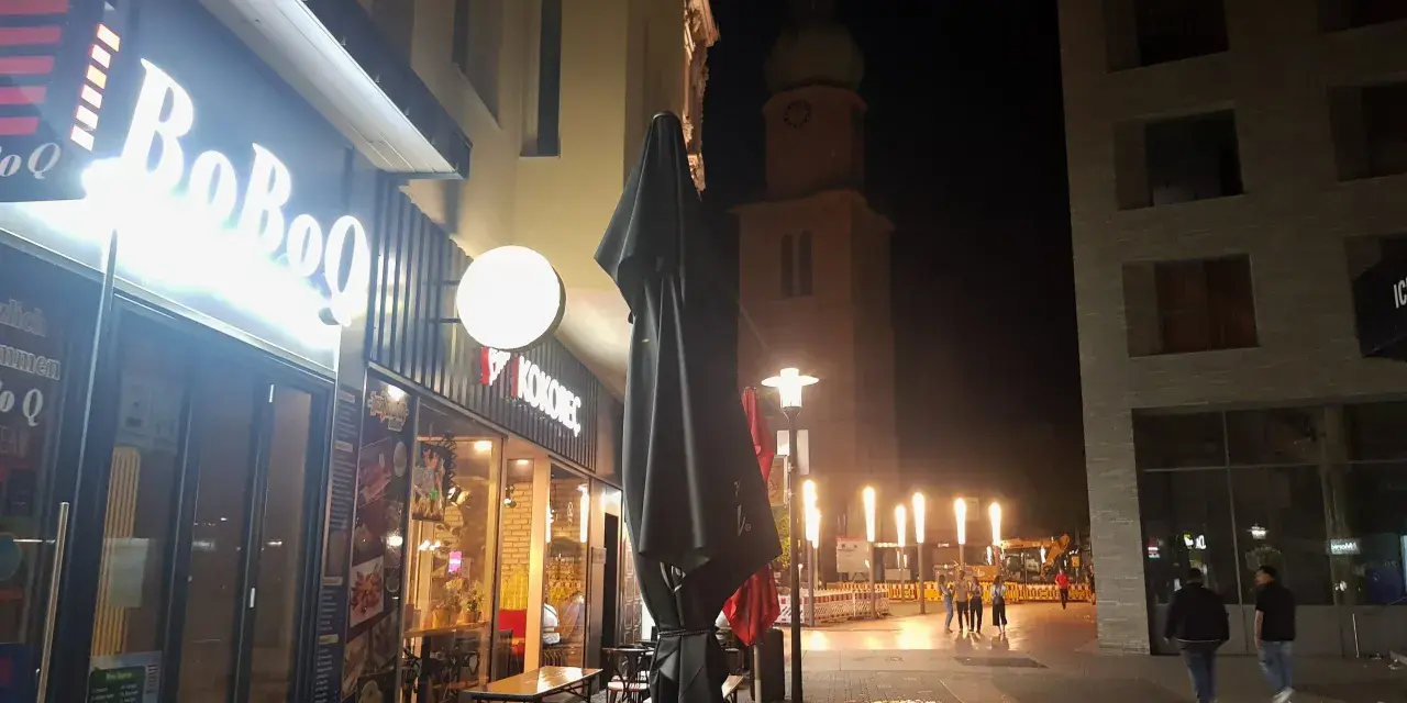 Ein beleuchtetes Restaurant "BoBoQ" und eine Kirche prägen das Bild von Dortmund bei Nacht. Menschen spazieren auf dem Gehweg, während Bauarbeiten im Hintergrund stattfinden.