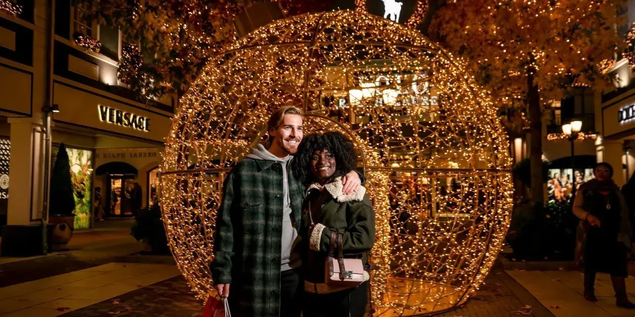 Ein glückliches Paar vor einer riesigen Lichterkugel auf dem weihnachtsmarkt roermond. Geschäfte wie Versace und Ralph Lauren sind im Hintergrund zu sehen.