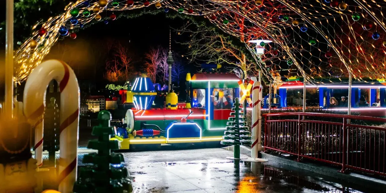 Ein bunter Zug fährt durch eine festlich beleuchtete Gasse im winterwonder legoland. Lichterketten und Kugeln schmücken den Weg.
