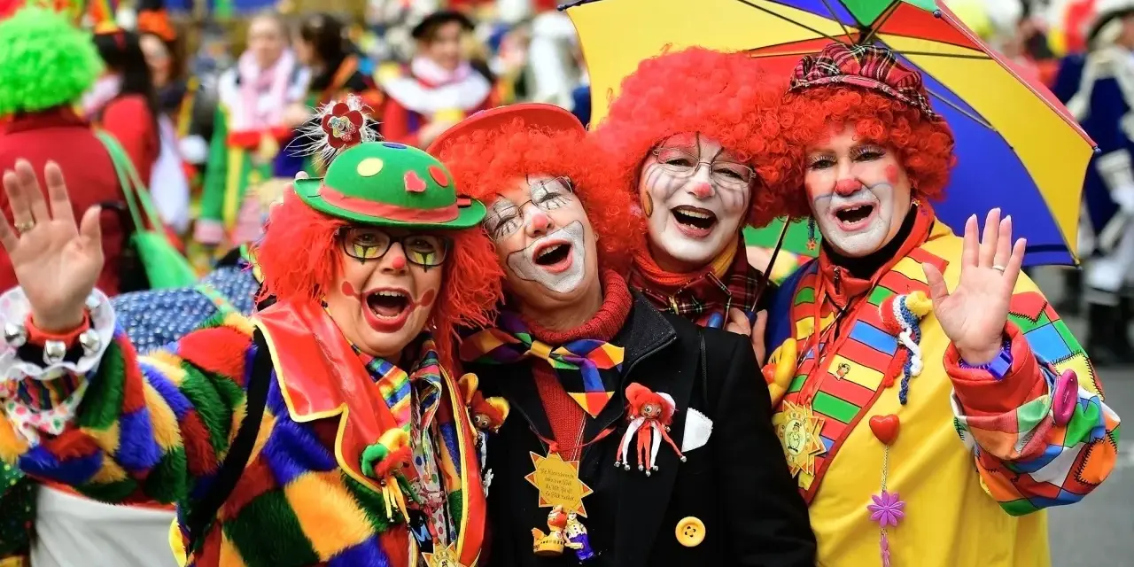 Vier Clowns mit roten Perücken und bunter Kleidung lachen und winken. Ein bunter Regenschirm ist über ihnen. Hier wird kein Karneval gefeiert, sondern ausgelassen gefeiert!