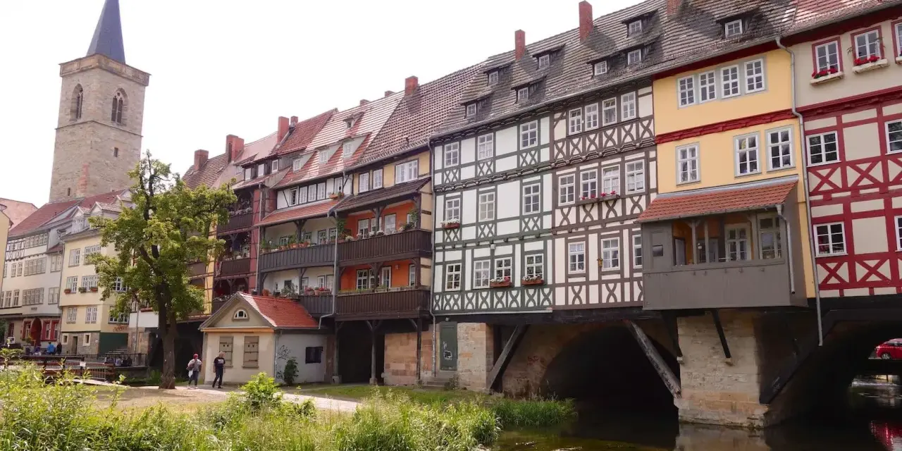 Fachwerkhäuser reihen sich am Wasser entlang, ein Kirchturm ragt empor. Ein idyllischer Anblick, um Erfurt entdecken zu gehen.