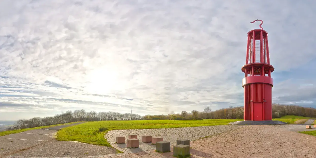 Rote Aussichtsturm auf der Halde Moers, umgeben von grünen Hügeln und einem weiten Himmel mit Wolken.
