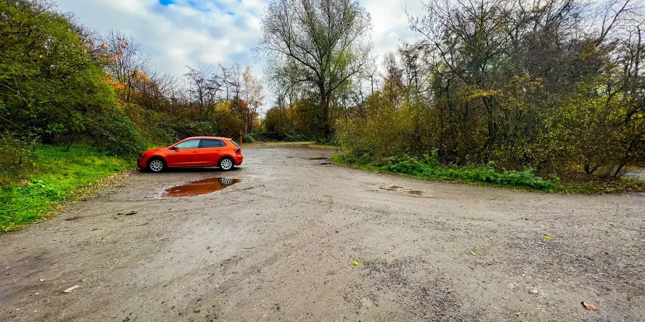 Ein orangefarbener VW Polo parkt auf einem matschigen Parkplatz am Rande eines Waldes. Die Halde Haniel Parken ist von Bäumen und Herbstlaub umgeben.