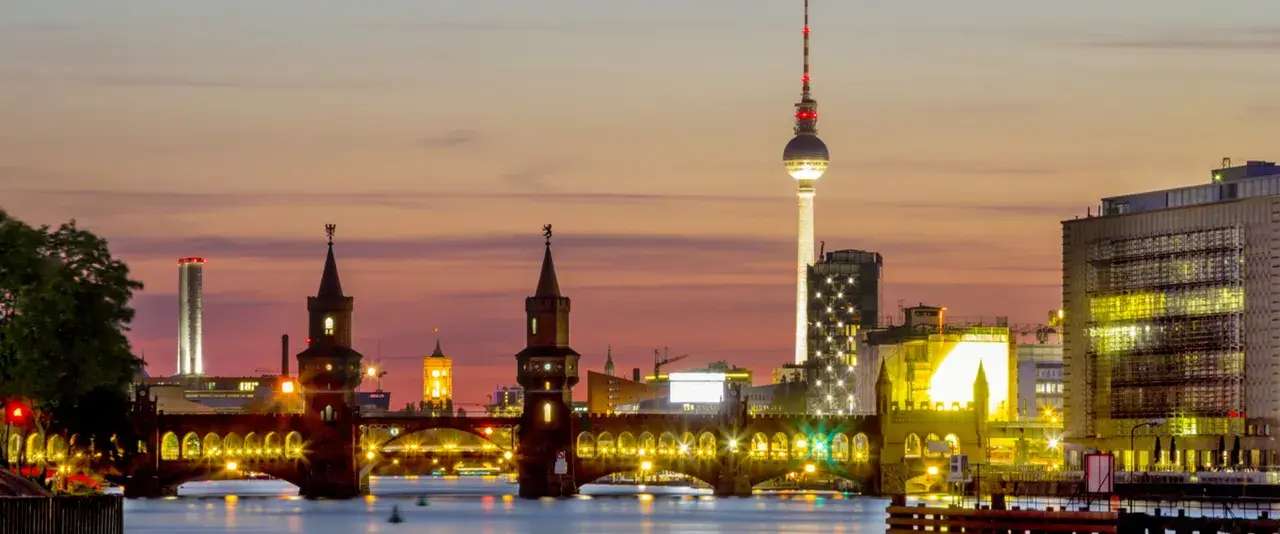 Berlin Kreuzberg bei Sonnenuntergang: Oberbaumbrücke, Fernsehturm und beleuchtete Gebäude spiegeln sich im Wasser.