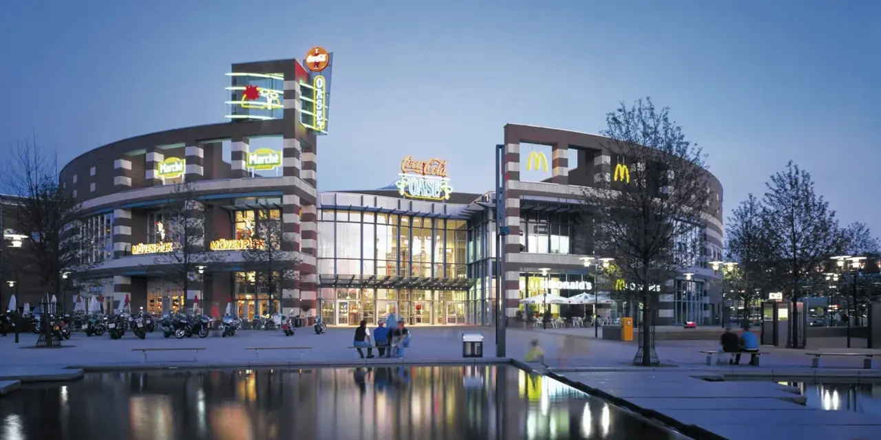 Abendstimmung am Centro Oberhausen. Die beleuchtete Fassade mit den Logos von McDonald's und Coca-Cola spiegelt sich im Wasser.