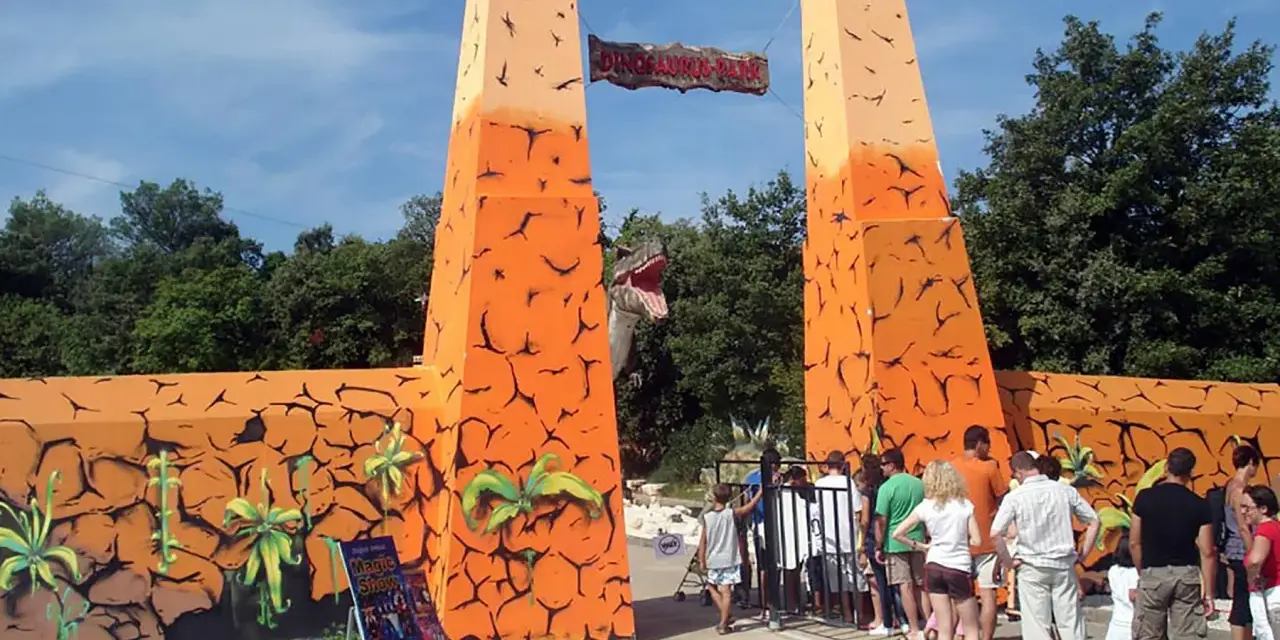 Eingang zum Dinosaurier-Park, einem Freizeitpark in Kroatien. Eine Gruppe von Besuchern steht vor dem Eingangstor.