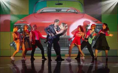 Tänzer in 50er-Jahre-Outfits vor einem roten Oldtimer-Cabriolet. Ein Paar tanzt im Zentrum, umgeben von anderen Darstellern. Ein lebendiger Moment aus Musicals Deutschland.