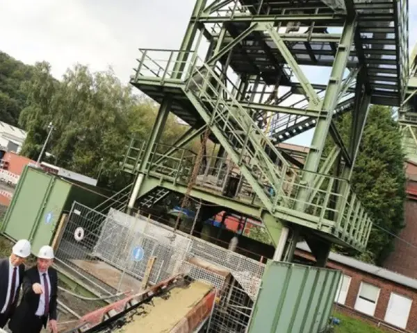 Drei Männer in Anzügen und Schutzhelmen inspizieren das Gelände der Zeche Bottrop. Ein Förderturm ragt in den Himmel.