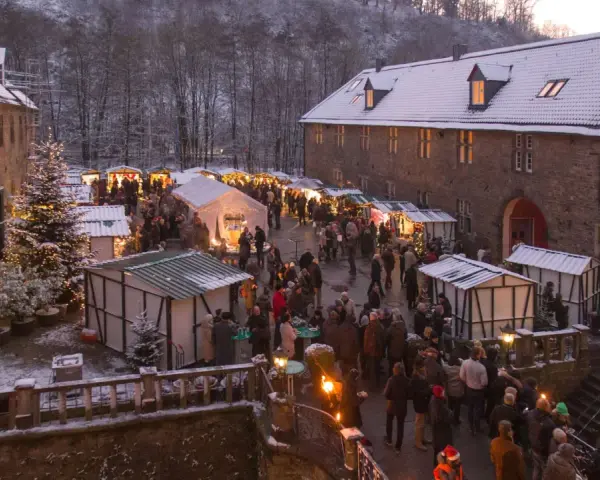 Schneebedeckter Weihnachtsmarkt in einer Burg, ein echter Geheimtipp NRW. Besucher schlendern zwischen festlich beleuchteten Buden.