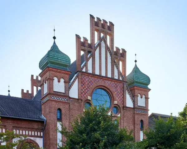Das imposante Gebäude im Stil des Zollern-Schlosses mit seinen grünen Kuppeln und roten Ziegeln ragt vor blauem Himmel auf.