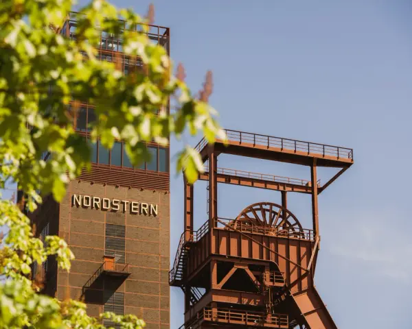 Industriearchitektur im Nordsternpark Gelsenkirchen: Ein markanter Förderturm und das Gebäude mit der Aufschrift "NORDSTERN" vor blauem Himmel.