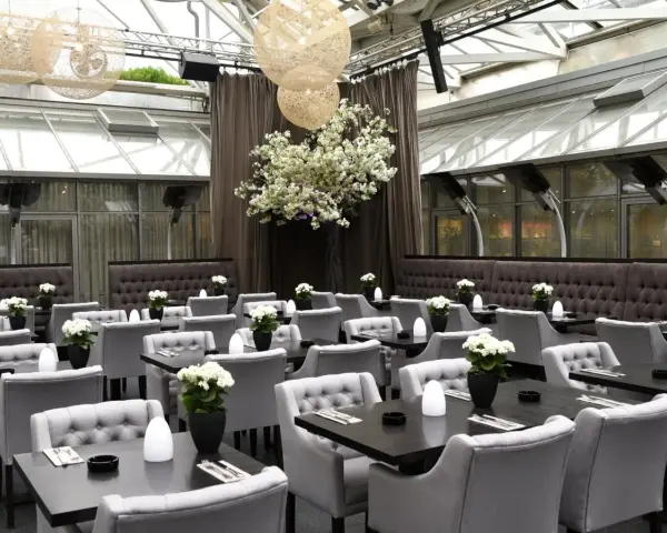 Elegantes Restaurant mit grauen Sitzmöbeln und dunklen Tischen, dekoriert mit weißen Blumen. Ein großer Kirschblütenbaum im Zentrum, beleuchtet von kugelförmigen Lampen. Perfekt für ein Rendezvous, wie ein Oscar-Moment im Apropos.