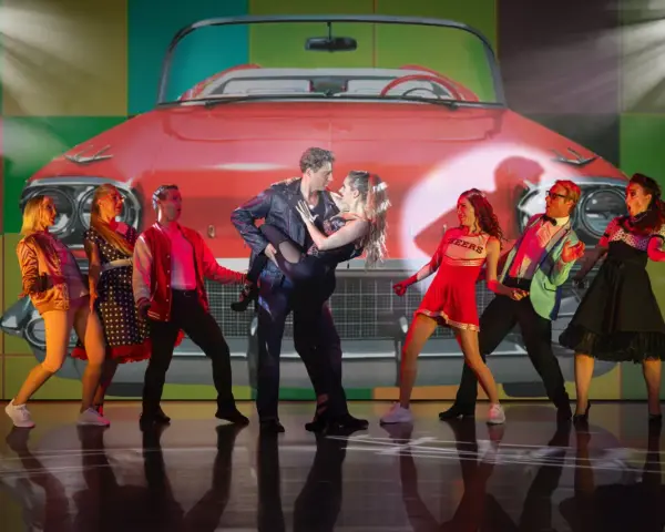 Tänzer in 50er-Jahre-Outfits vor einem roten Oldtimer-Cabriolet. Ein Paar tanzt im Zentrum, umgeben von anderen Darstellern. Ein lebendiger Moment aus Musicals Deutschland.