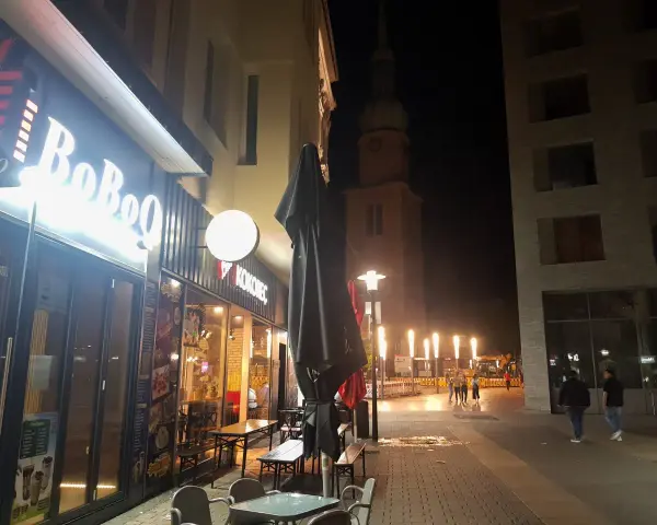 Ein beleuchtetes Restaurant "BoBoQ" und eine Kirche prägen das Bild von Dortmund bei Nacht. Menschen spazieren auf dem Gehweg, während Bauarbeiten im Hintergrund stattfinden.