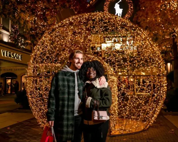 Ein glückliches Paar vor einer riesigen Lichterkugel auf dem weihnachtsmarkt roermond. Geschäfte wie Versace und Ralph Lauren sind im Hintergrund zu sehen.