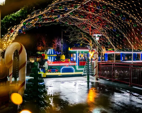 Ein bunter Zug fährt durch eine festlich beleuchtete Gasse im winterwonder legoland. Lichterketten und Kugeln schmücken den Weg.