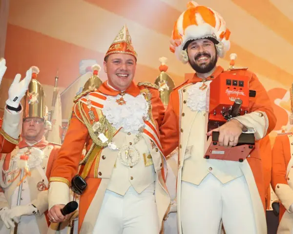 Narren in bunten Kostümen feiern jecke Tage. Ein Mann hält eine Kaffeemaschine, die anderen tragen prunkvolle Uniformen und Federhüte.