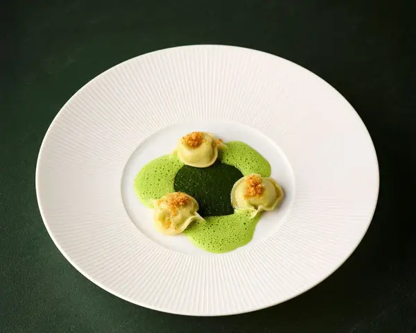 Ammolite speisekarte: Drei grüne Ravioli mit goldbrauner Kruste auf einer leuchtend grünen Sauce, angerichtet auf einem weißen Teller.