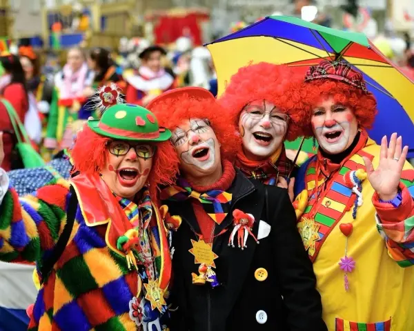 Vier Clowns mit roten Perücken und bunter Kleidung lachen und winken. Ein bunter Regenschirm ist über ihnen. Hier wird kein Karneval gefeiert, sondern ausgelassen gefeiert!