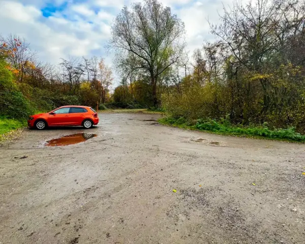 Ein orangefarbener VW Polo parkt auf einem matschigen Parkplatz am Rande eines Waldes. Die Halde Haniel Parken ist von Bäumen und Herbstlaub umgeben.