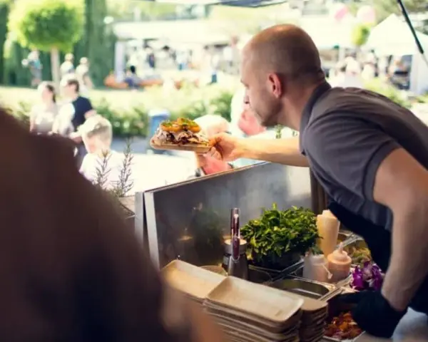 Mann reicht ein Gericht auf einem Holzbrett bei einem Street Food Festival. Die Preise sind hier fair für die leckere Auswahl.