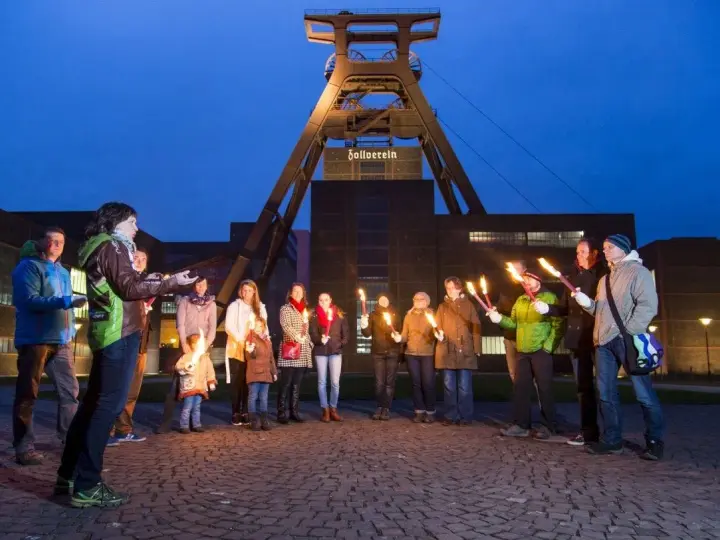 Eine Gruppe von Menschen hält nachts Fackeln vor dem Förderturm der Zeche Zollverein.