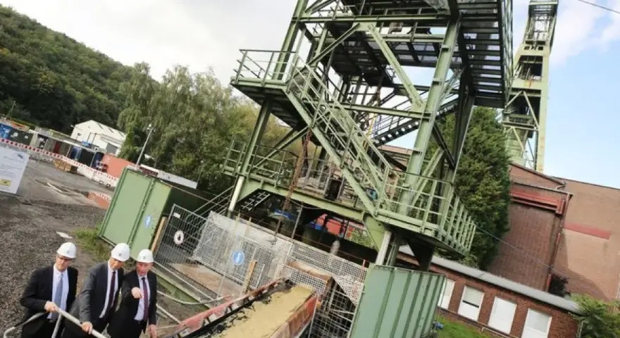 Drei Männer in Anzügen und Schutzhelmen inspizieren das Gelände der Zeche Bottrop. Ein Förderturm ragt in den Himmel.