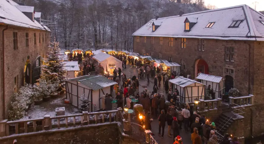 Schneebedeckter Weihnachtsmarkt in einer Burg, ein echter Geheimtipp NRW. Besucher schlendern zwischen festlich beleuchteten Buden.