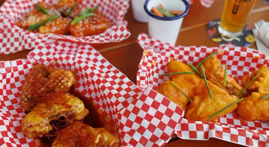 Leckere Cosmo Wings in roten Körbchen, garniert mit Chilis und Schnittlauch. Dazu ein Bier und kleine Beilagen.