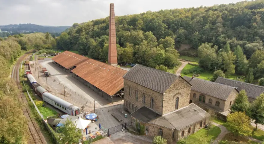 Alte Zechengebäude der Zeche Nachtigall mit einem langen roten Dach, einem hohen Backsteinschornstein und einem historischen Zug.