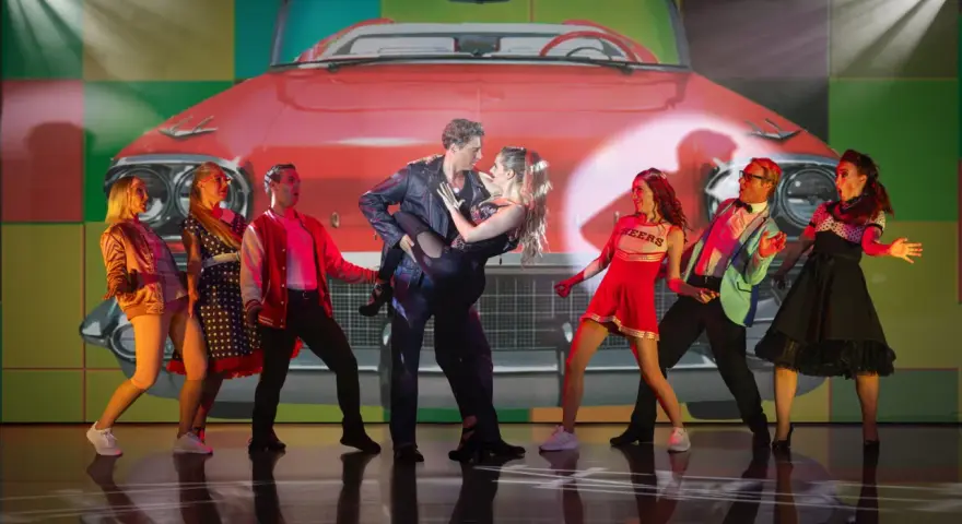 Tänzer in 50er-Jahre-Outfits vor einem roten Oldtimer-Cabriolet. Ein Paar tanzt im Zentrum, umgeben von anderen Darstellern. Ein lebendiger Moment aus Musicals Deutschland.