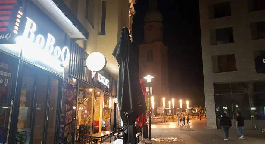 Ein beleuchtetes Restaurant "BoBoQ" und eine Kirche prägen das Bild von Dortmund bei Nacht. Menschen spazieren auf dem Gehweg, während Bauarbeiten im Hintergrund stattfinden.