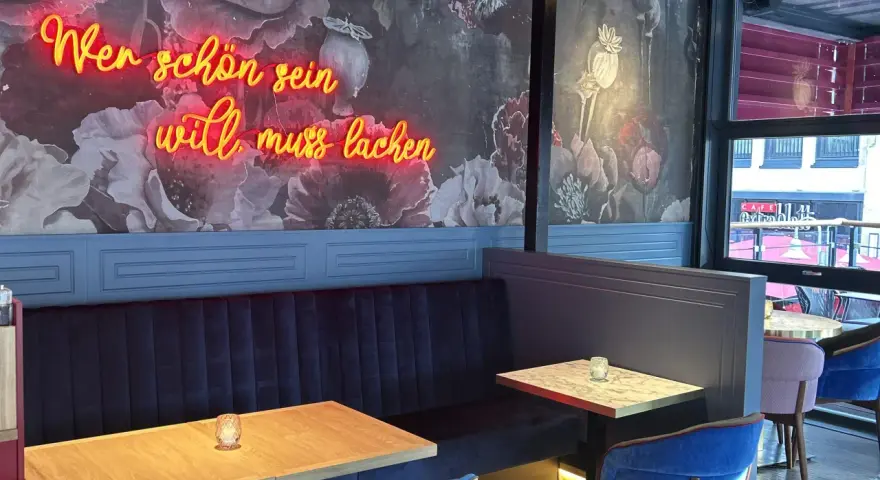 Wilma Wunder: Neonlicht über blauer Bank und floraler Tapete. Wer schön sein will, muss lachen.